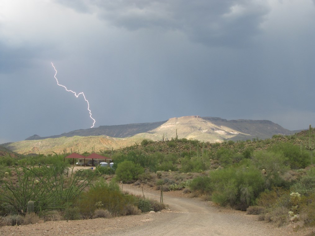 Lightning bolt striking near a desert mesa under dark storm clouds, with saguaro cacti and a small picnic shelter in the foreground.