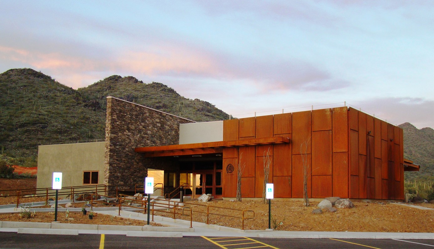 View of the Cave Creek Regional Park Nature Center with a blue sky and mountain in the backgroun. Facility is made of cement, patina panels, and natural rock.