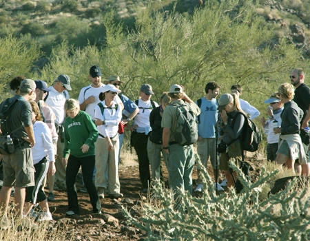 A group of people gather on the Elephant Mountain Trail for a guided hike with the park ranger in the Spur Cross Ranch Conservation Area, surrounded by desert vegetation and rocky terrain.