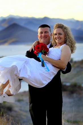 Bride and groom posing on a desert hillside at the Desert Outdoor Center, with the groom holding the bride and Lake Pleasant and mountain views in the background.