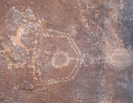 Petroglyph carved into desert rock showing a circular design with a central dot and surrounding lines, likely created by Indigenous peoples.