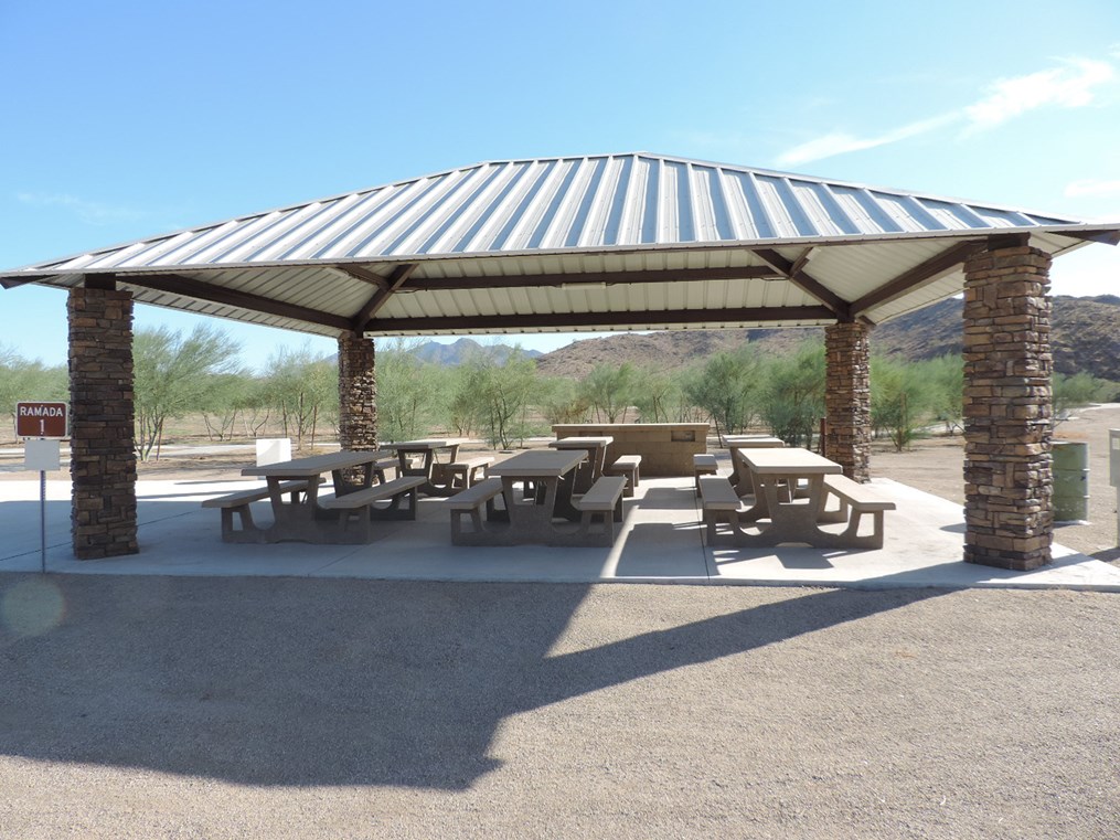 Ramada 1 picnic shelter with metal roof, picnic tables on a cement pad, nearby parking, and accessible pathway.