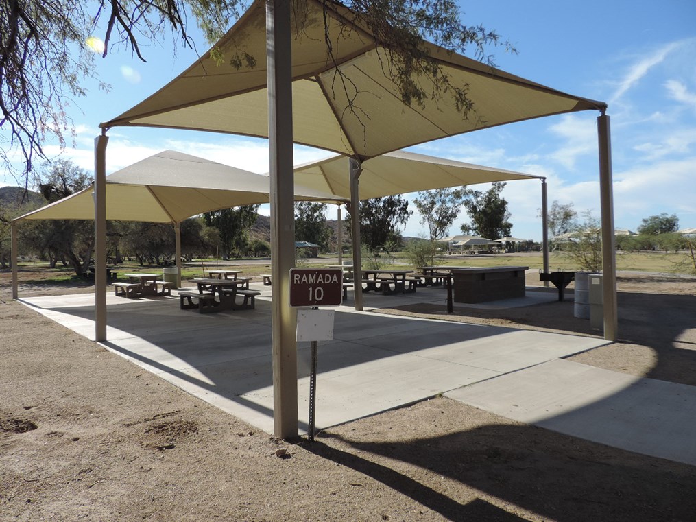 Ramada 10 shade-covered picnic area with tables on a cement pad, accessible walkway, and nearby open field.