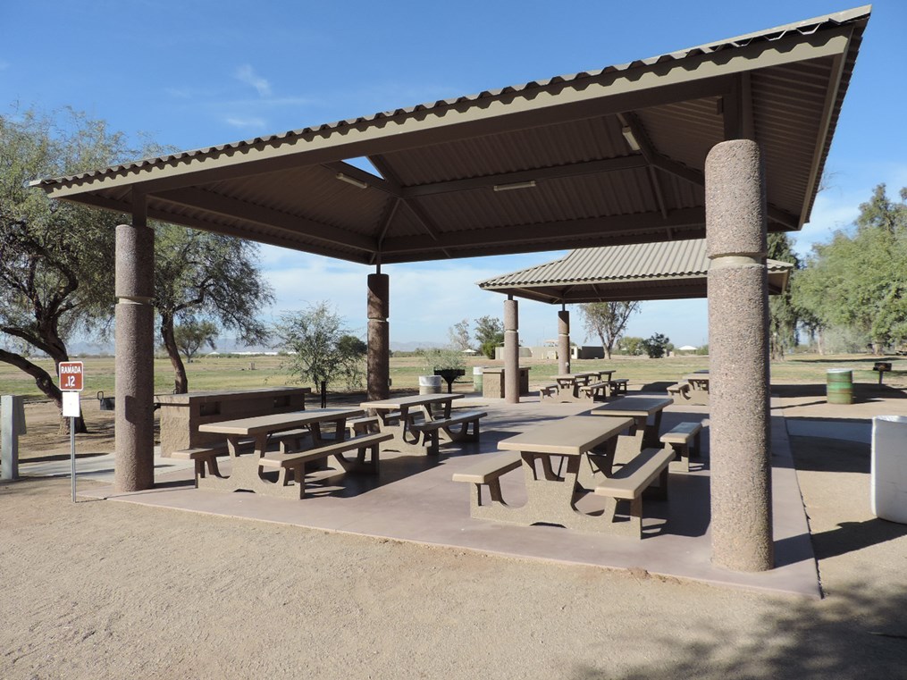 Ramada 12 picnic shelter with metal roof, picnic tables on a cement pad, accessible sidewalk connection, and nearby open field.