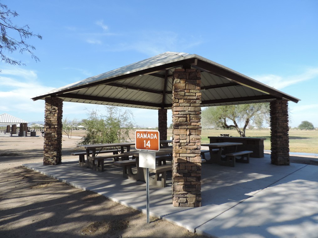 Ramada 14 picnic shelter with metal roof, picnic tables on a cement pad, accessible path, and nearby grassy area.