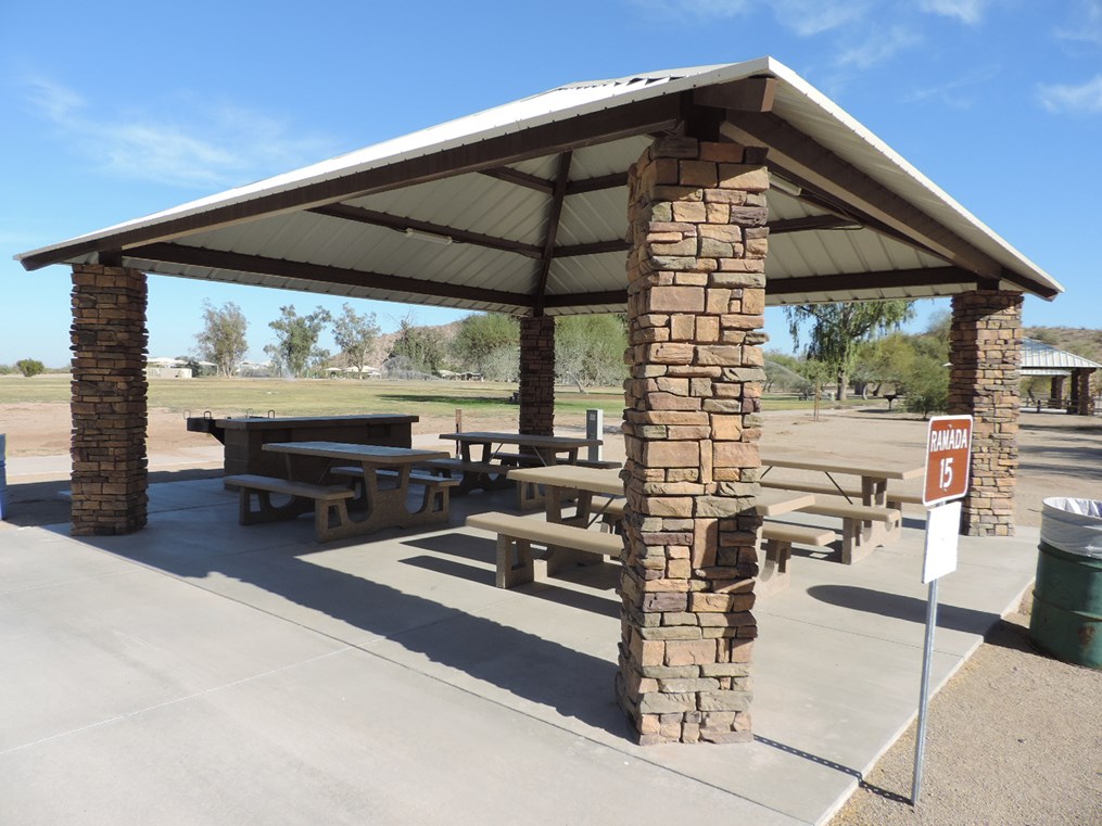 Ramada 15 picnic shelter with metal roof, picnic tables on a cement slab, accessible walkway, and open park views.