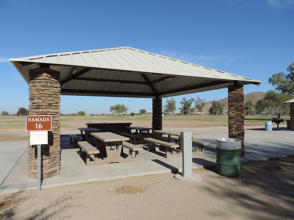 Ramada 16 picnic shelter with metal roof, picnic tables on a cement pad, accessible sidewalk connection, and open desert landscape.