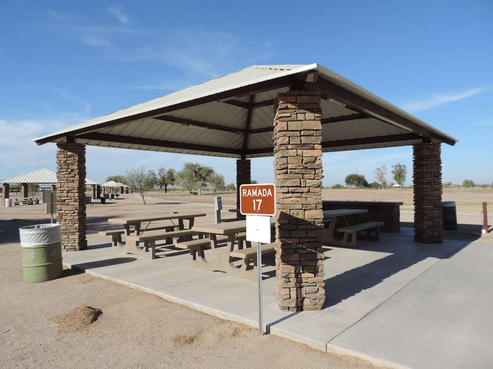 Ramada 17 picnic shelter with metal roof, picnic tables on a cement slab, accessible walkway, and nearby park lawn.