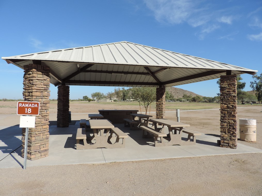 Ramada 18 picnic shelter with metal roof, picnic tables on a cement pad, accessible path, and open field backdrop.