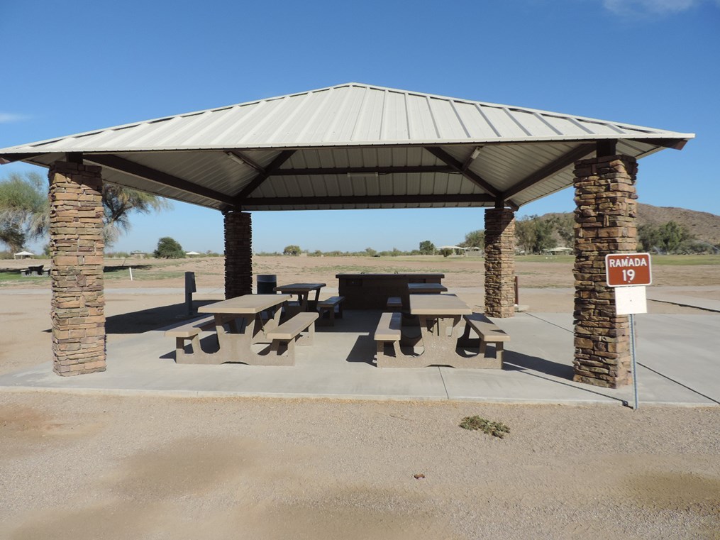 Ramada 19 picnic shelter with metal roof, picnic tables on a cement slab, accessible sidewalk connection, and desert park setting.