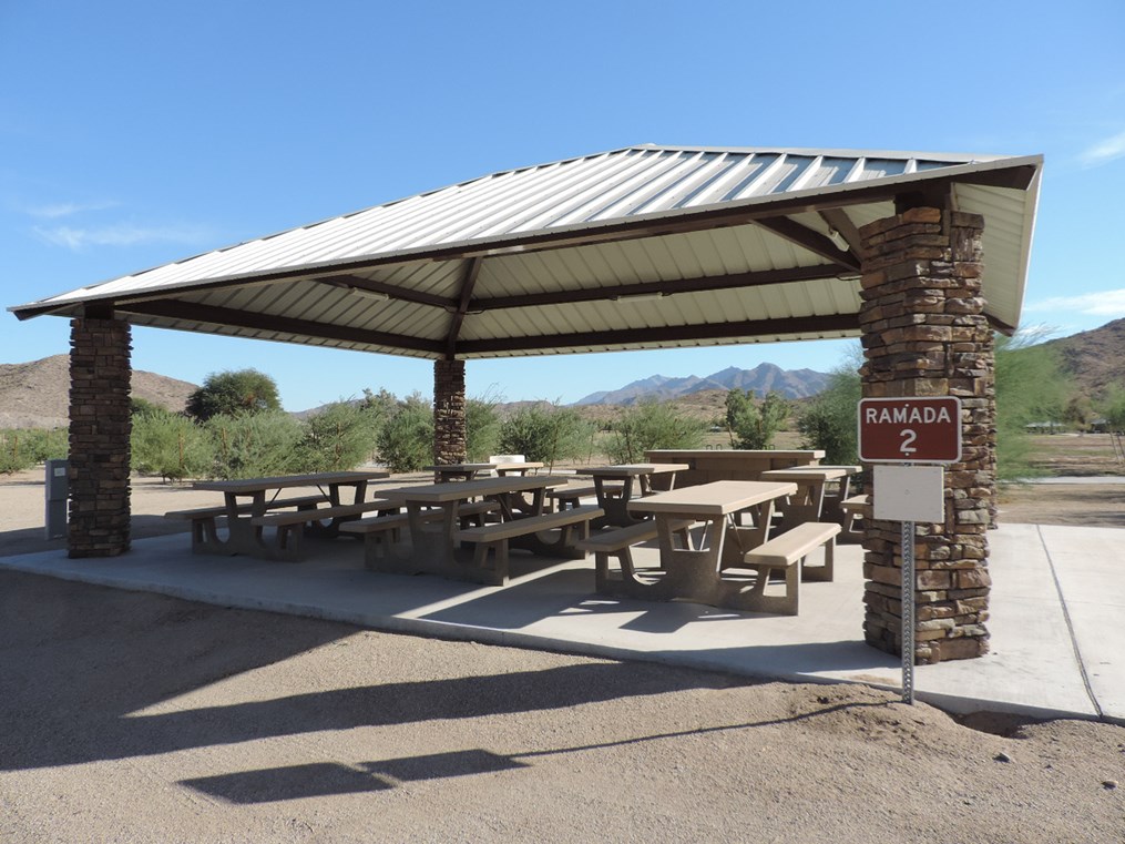 Ramada 2 picnic shelter with metal roof, picnic tables on a cement pad, nearby parking, and accessible sidewalk connection.
