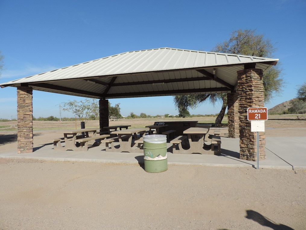Ramada 21 picnic shelter with metal roof, picnic tables on a cement slab, accessible path, and surrounding park landscape.
