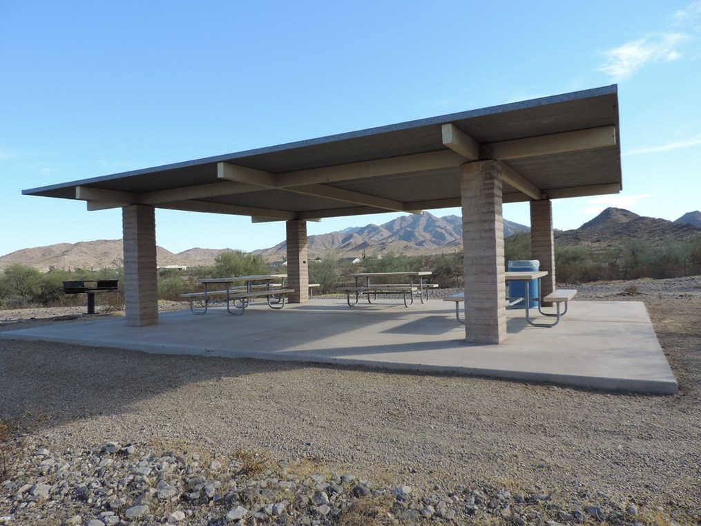 Ramada 27 picnic shelter with metal roof and picnic tables on a cement pad in a desert setting.