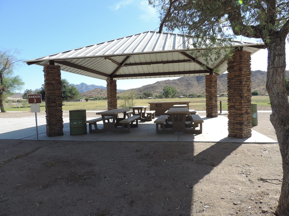 Ramada 3 picnic shelter with metal roof, picnic tables on a cement slab, accessible path, and open park views.