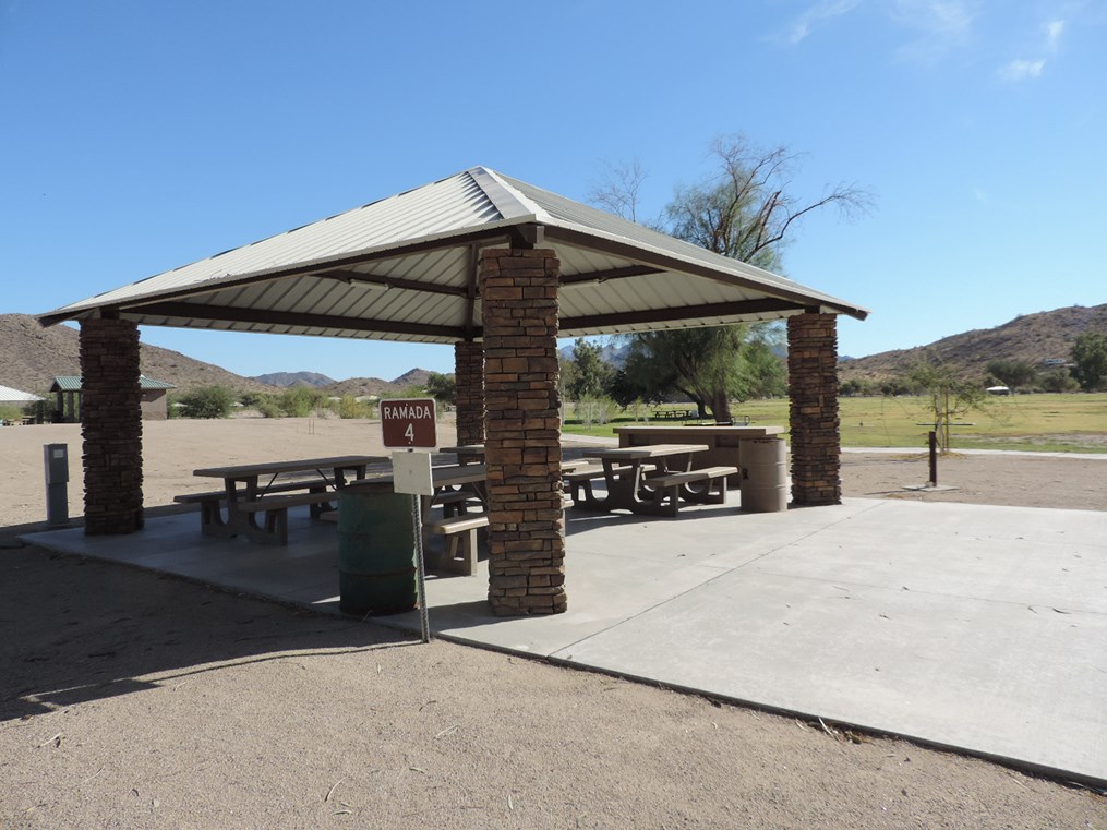 Ramada 4 picnic shelter with metal roof, picnic tables on a cement pad, accessible walkway, and nearby grassy area.