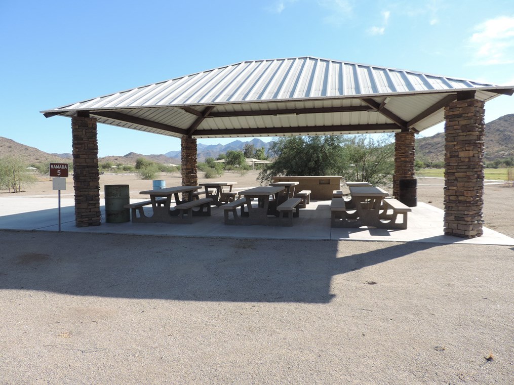 Ramada 5 picnic shelter with metal roof, picnic tables on a cement pad, accessible sidewalk, and mountain backdrop.