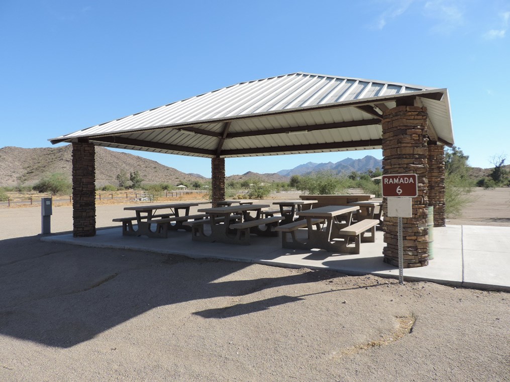 Ramada 6 picnic shelter with metal roof, picnic tables on a cement slab, accessible path, and desert surroundings.
