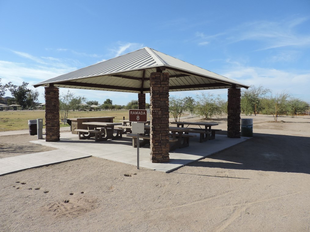 Ramada 8 picnic shelter with metal roof, picnic tables on a cement pad, accessible walkway, and open park setting.