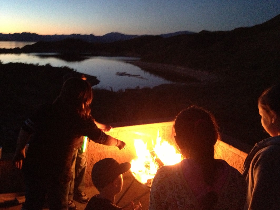 Group of people gathered around a campfire at dusk overlooking Lake Pleasant, with mountains and water visible in the background.