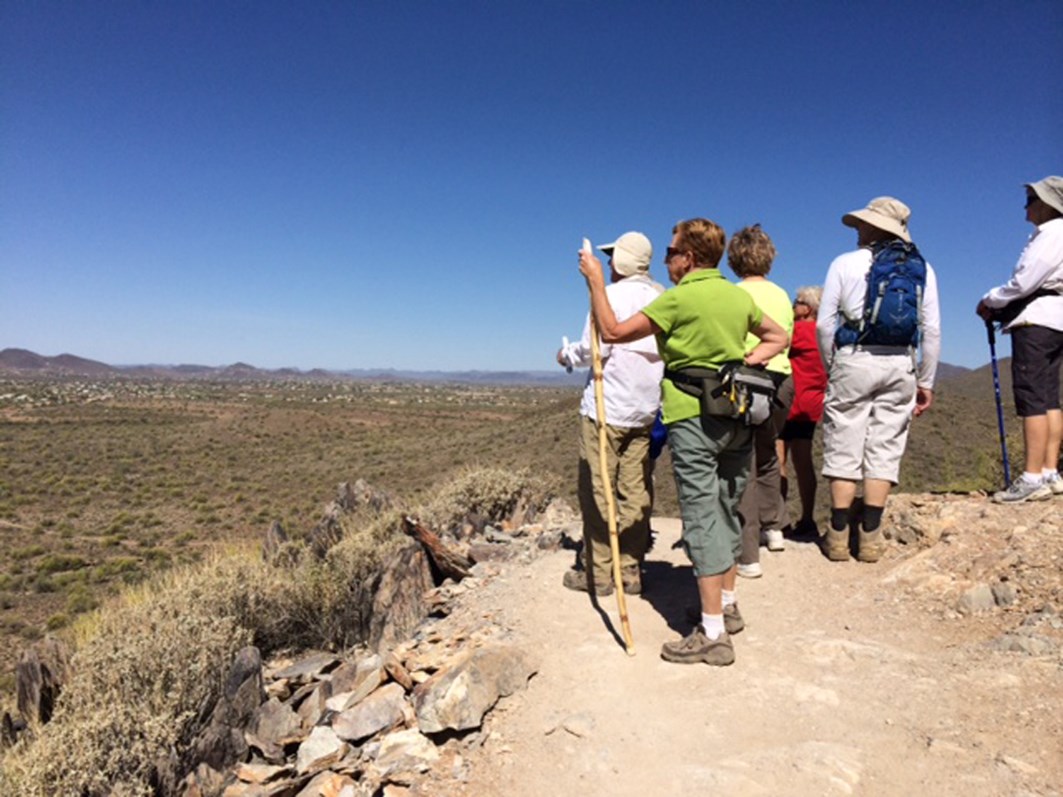 A group of hikers standing on a rocky desert trail, looking out over the desert landscape beneath a clear blue sky. 