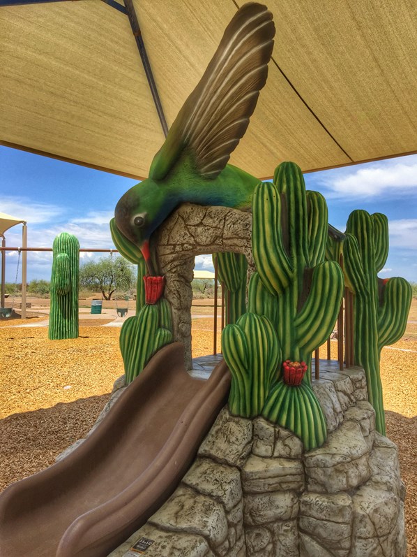 Hummingbird-themed playground slide at Estrella Mountain Regional Park, featuring a large green hummingbird sculpture above a brown slide surrounded by saguaro cactus designs and shade canopy.