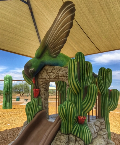 Hummingbird-themed playground slide at Estrella Mountain Regional Park, featuring a large green hummingbird sculpture above a brown slide surrounded by saguaro cactus designs and shade canopy.