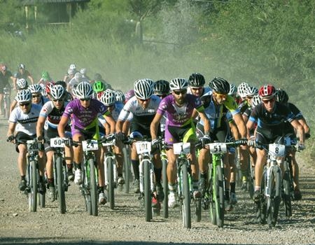 A group of cyclists races on a dirt trail during a mountain bike event at Spur Cross Ranch Conservation Area, kicking up dust as they ride closely together.