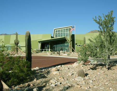 Exterior view of the White Tank Mountain Regional Park Nature Center, featuring a modern green building that is also home to a Maricopa County library and surrounded by desert landscaping and saguaro cacti under a clear blue sky.