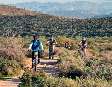 Group of mountain bikers riding along a desert trail with scenic mountain views in the background.