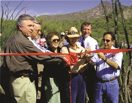 A group of people cut a red ribbon during a ceremony in a desert setting, with mountains and vegetation in the background in the Spur Cross Ranch Conservation Area.